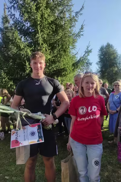 Młody mężczyzna z nagrodą i kobieta w czerwonej koszulce z logo, na zewnątrz w słońcu.