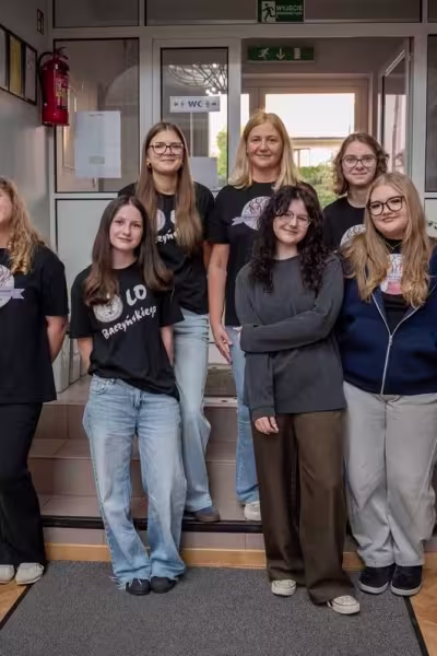 Grupa dziewięciu osób, głównie młodych kobiet, stoi w szkolnym korytarzu.