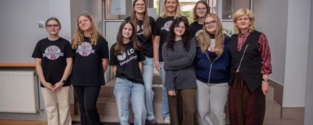 Grupa dziewięciu osób, głównie młodych kobiet, stoi w szkolnym korytarzu.